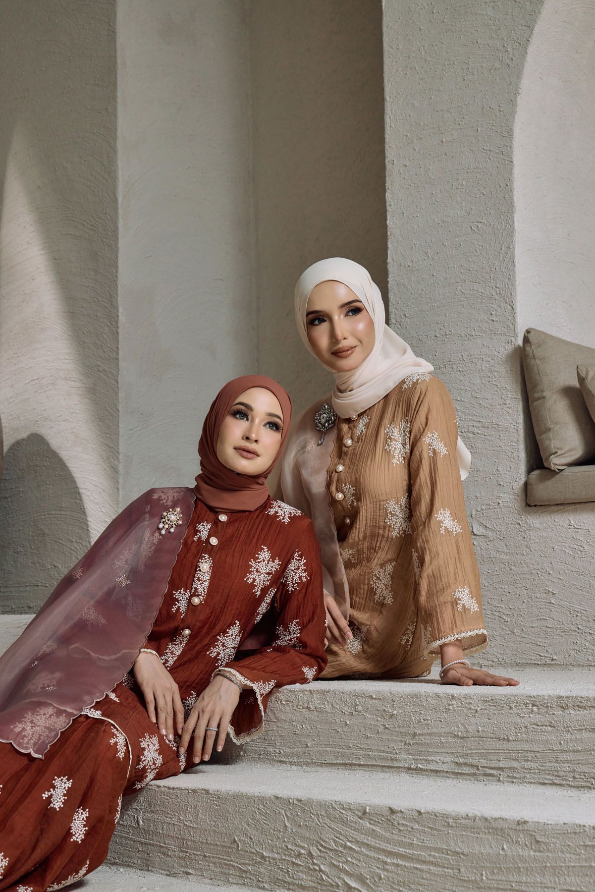Two women in hijabs wearing embroidered baju kurung in rust red and tan brown with floral lace details.