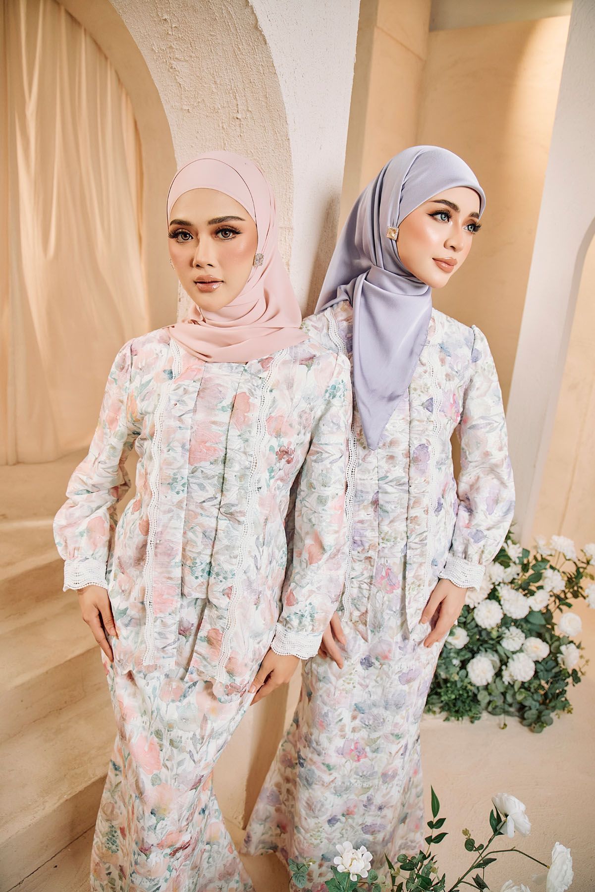 Two women in matching floral baju kurung with pink and lavender hijabs pose together.