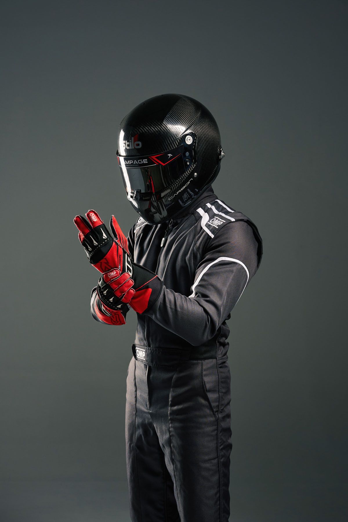 Racing driver in black suit and red gloves adjusting gear, wearing carbon fiber helmet against gray background.