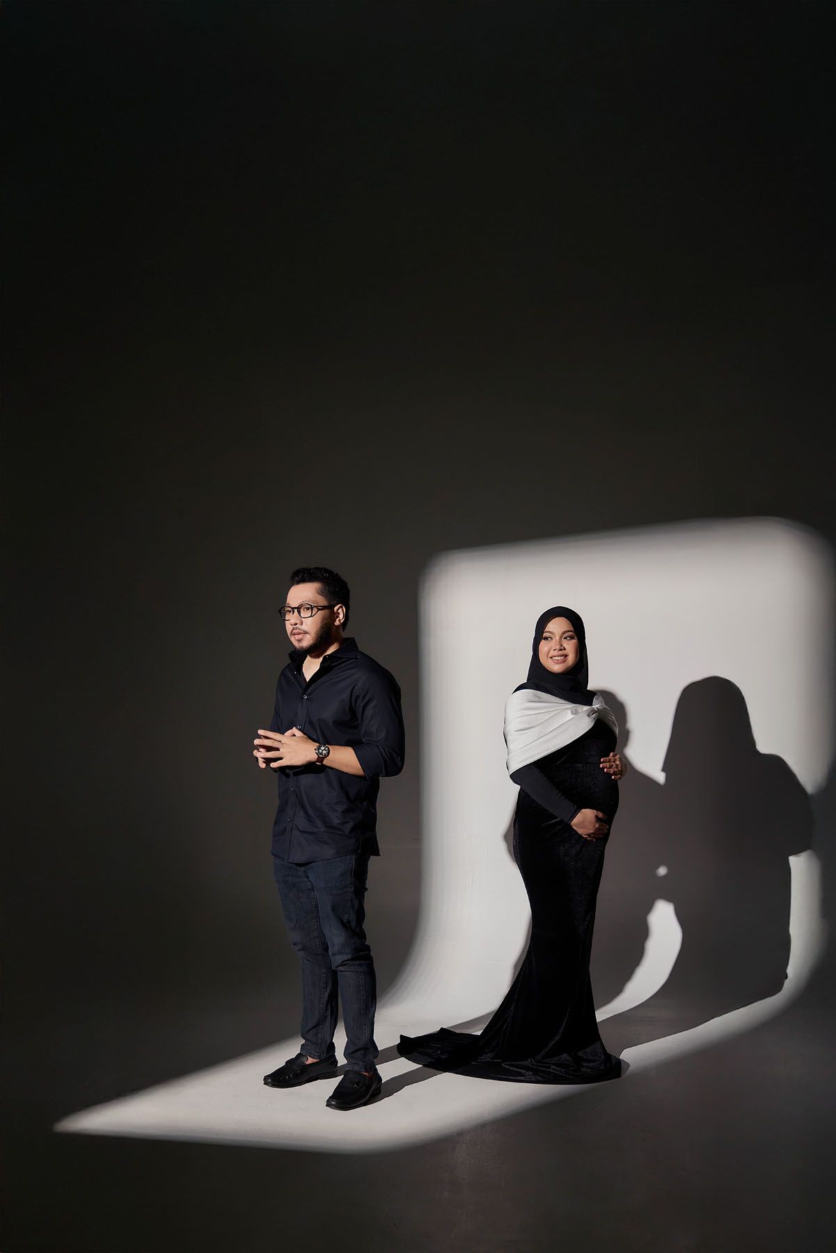 Couple posing in spotlight, man in navy outfit beside woman in black hijab gown with shadows.