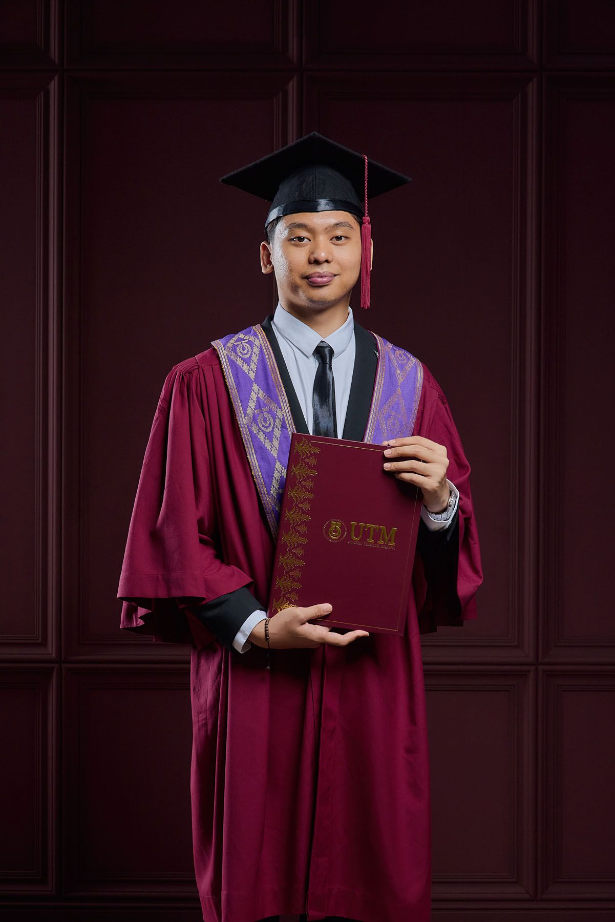 Smiling male graduate in maroon UTM academic robe presenting university diploma book to camera.
