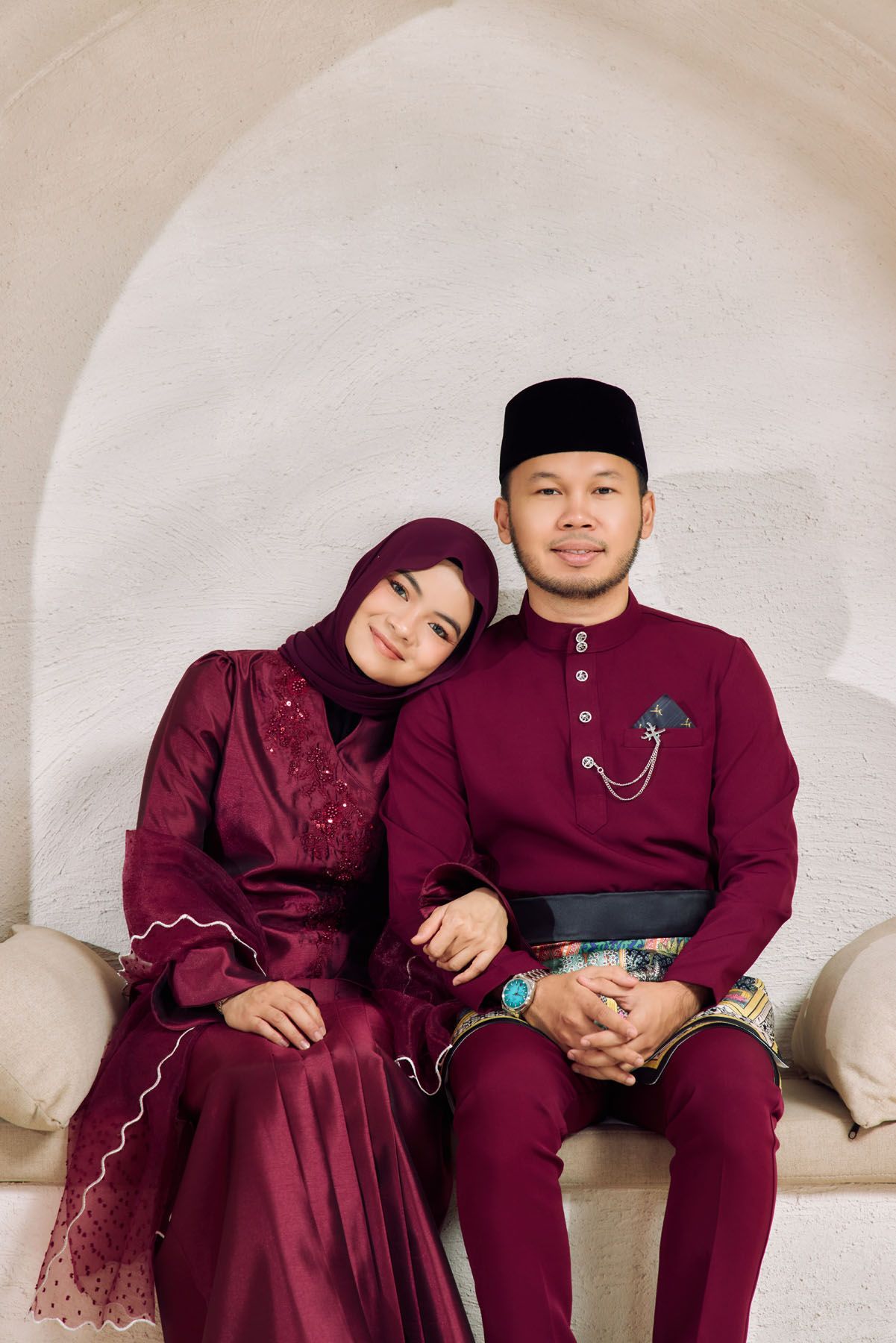 A Malaysian couple in matching deep red traditional Baju Melayu and Baju Kurung sitting together under a white arch.