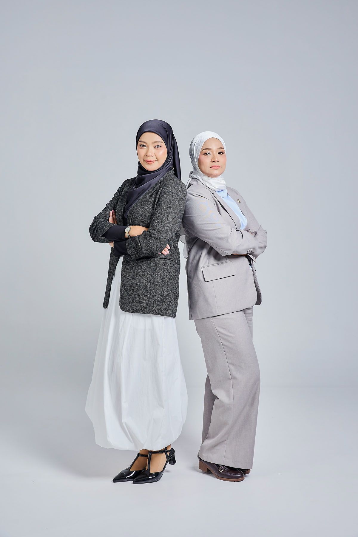 Two Muslim women in blazers and hijabs standing back-to-back with arms crossed on gray background.