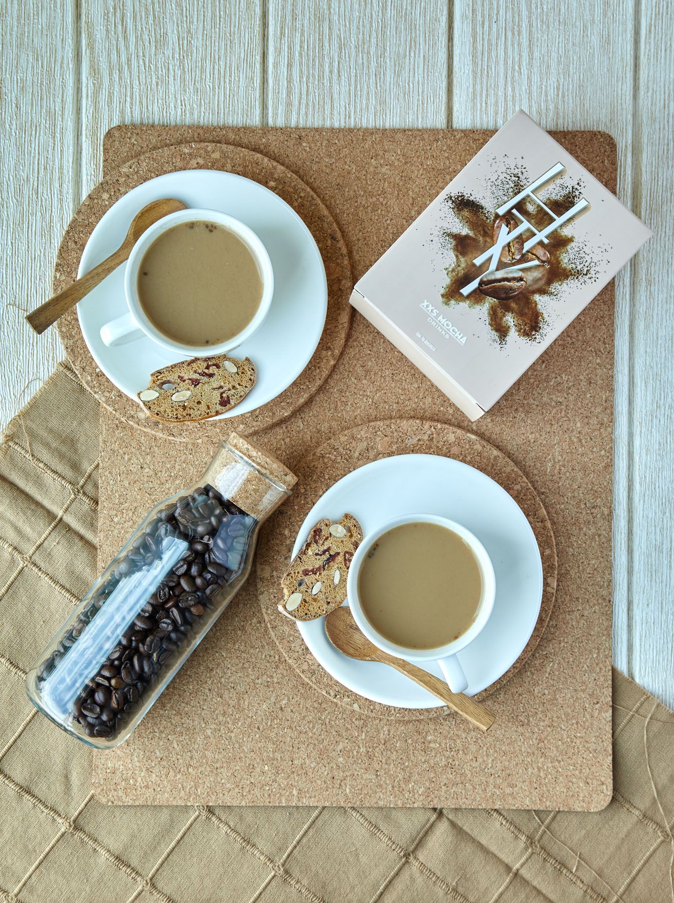 Top view of two coffee cups with biscotti on cork coasters beside coffee beans jar on beige surface.