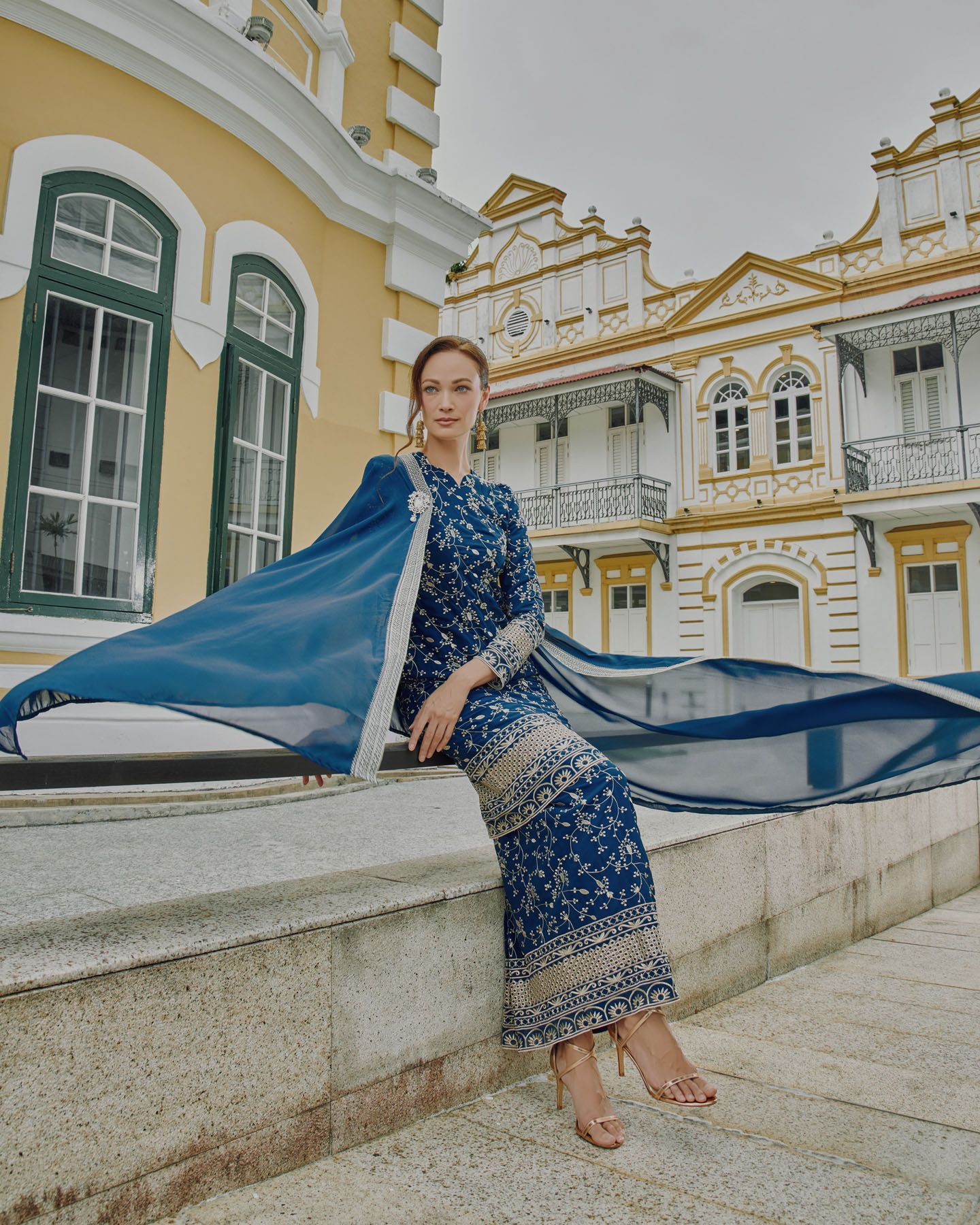 Woman in blue embroidered baju kurung with cape sits on a ledge before a colonial building.