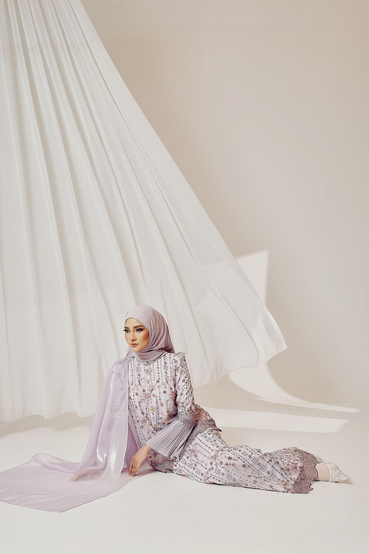 Muslim woman in lavender embellished gown and matching hijab seated gracefully against white draped fabric backdrop.
