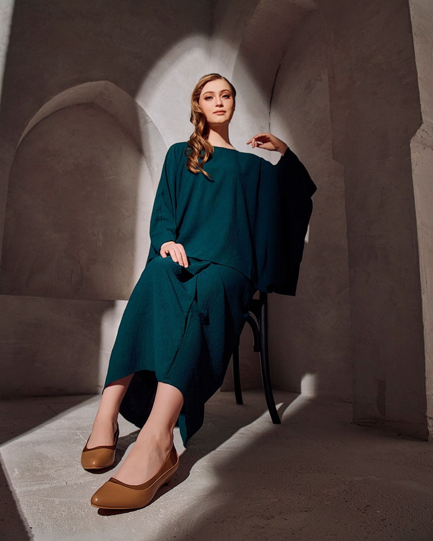 Woman in teal dress seated on chair wearing tan flats in arched stone setting.