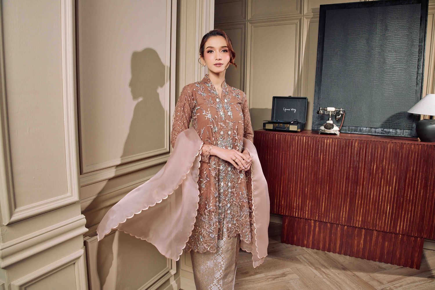 Woman in a shimmering beige embroidered kebaya with matching pants and dupatta scarf in elegant interior.
