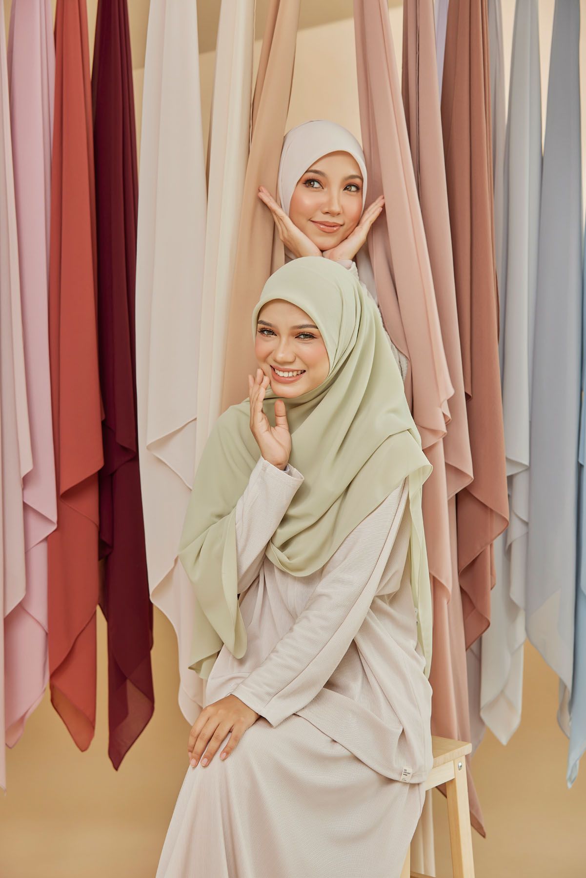Two Muslim women in light green and white hijabs posing before colorful hijab fabric display.