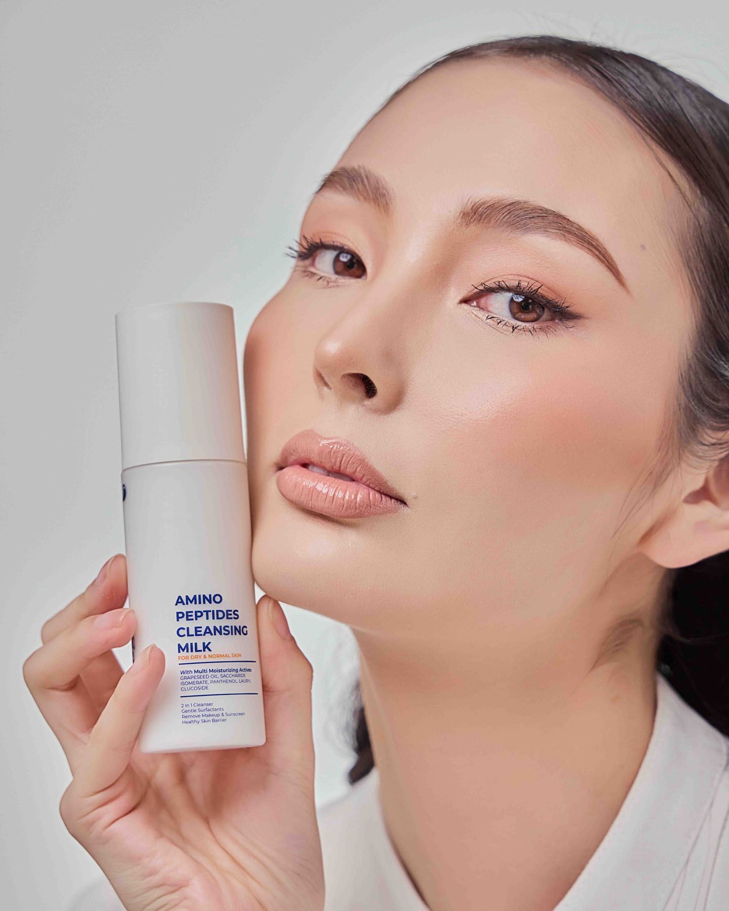 Asian woman holds Amino Peptides Cleansing Milk skincare product against white background.