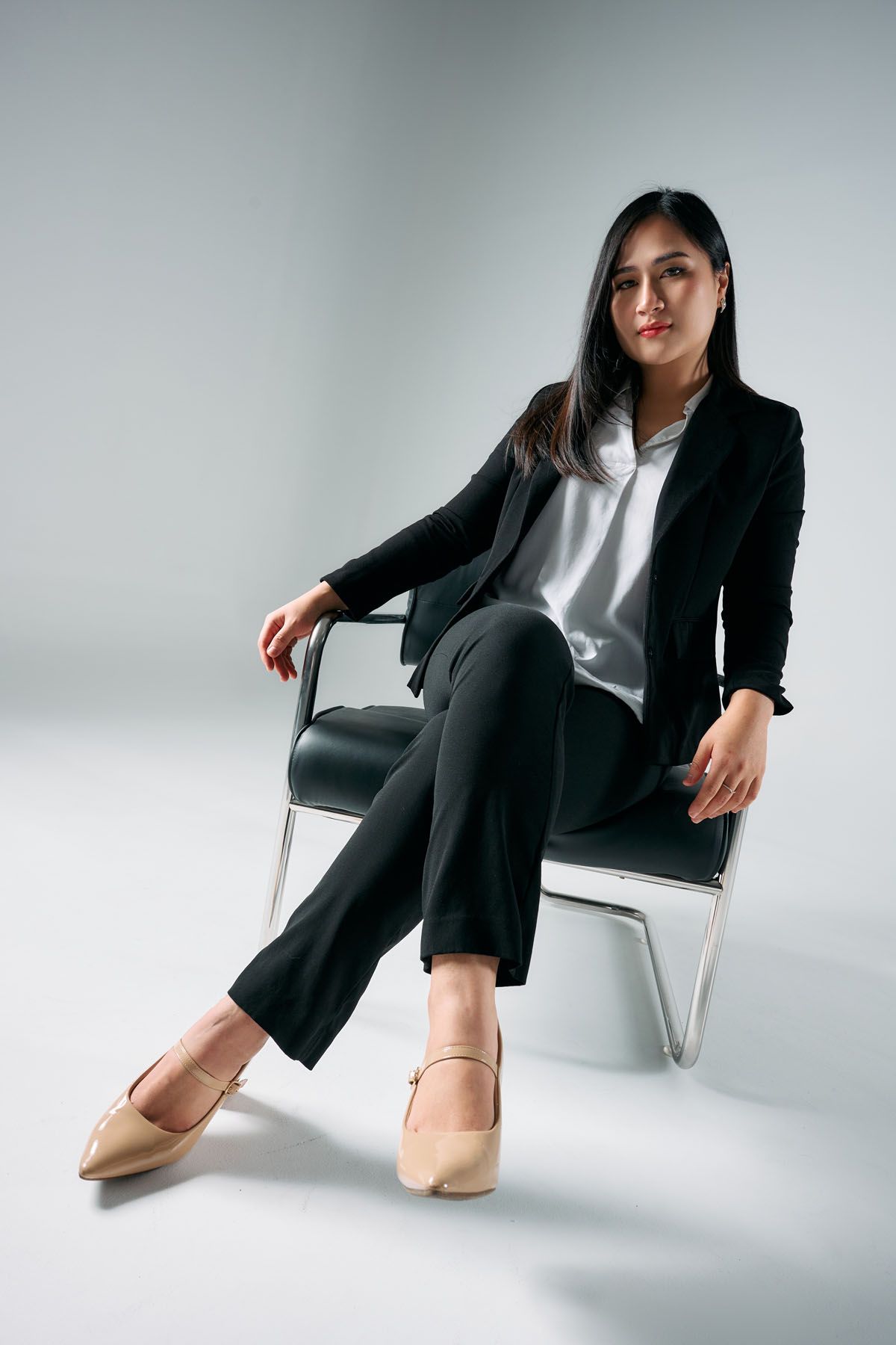 Professional woman in black suit sitting confidently on a chair against a white background.