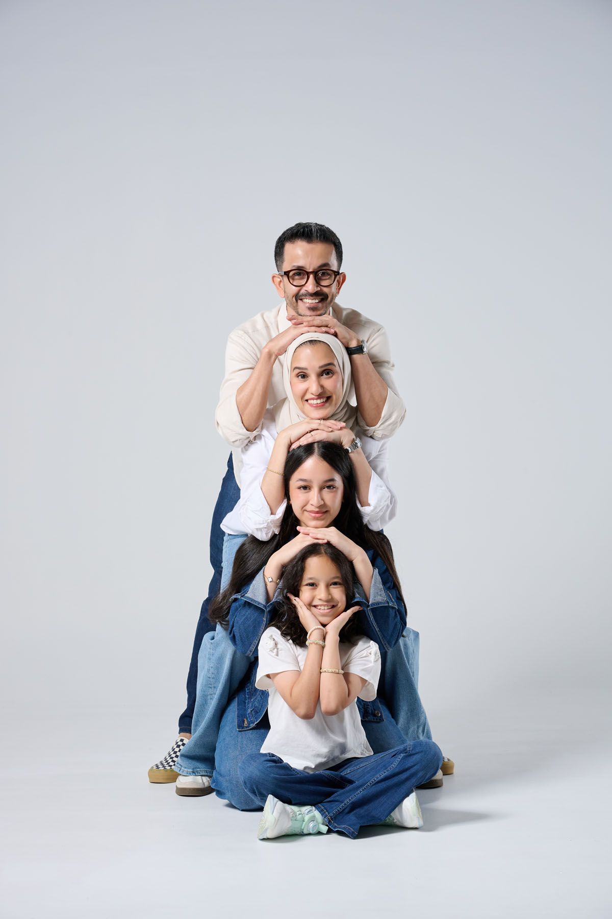 Happy Muslim family of four posing in a stacked formation, wearing casual white tops and jeans on grey background.