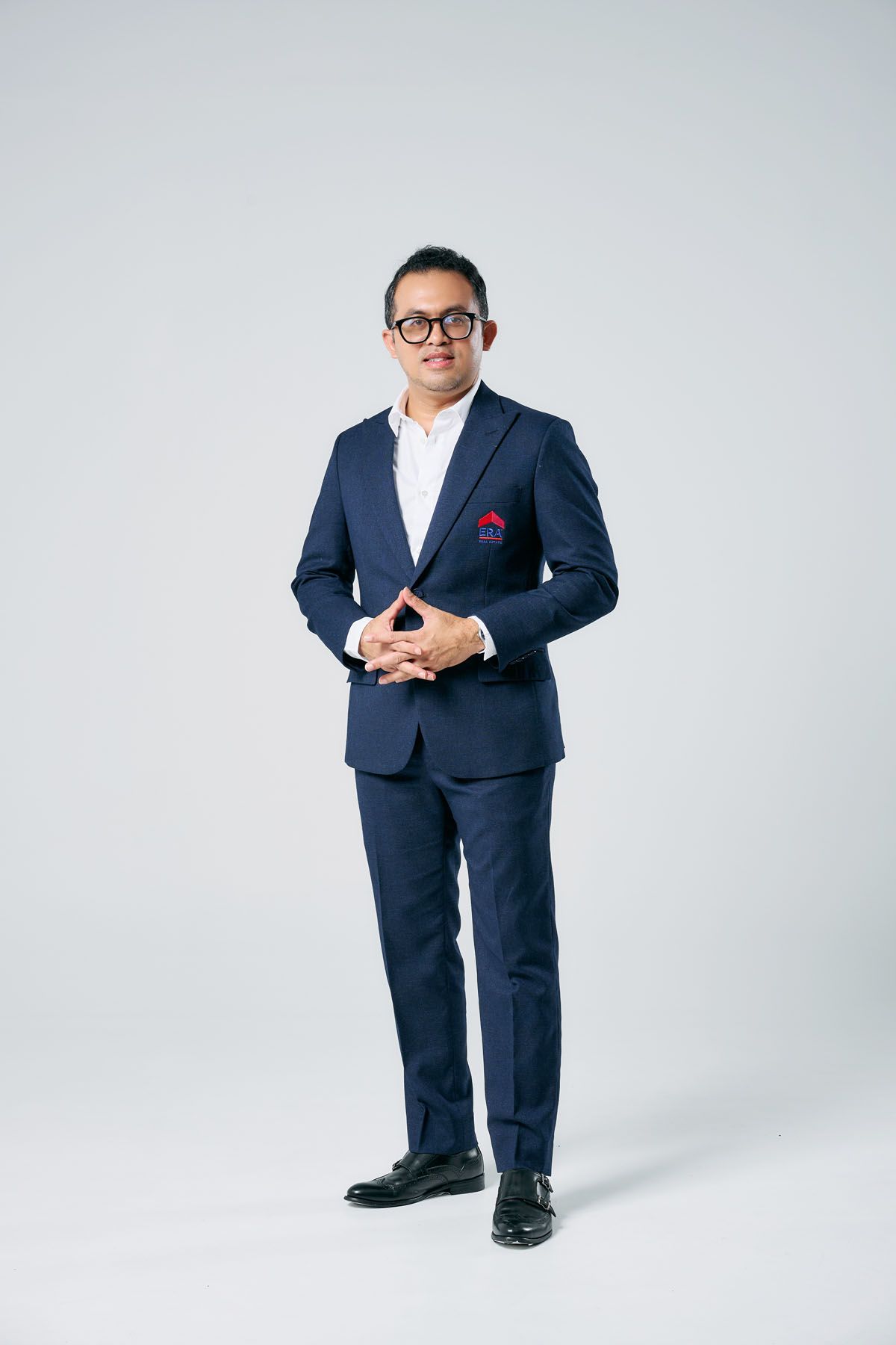 Professional man in navy blue suit with red pocket square and glasses, posing confidently against white background.