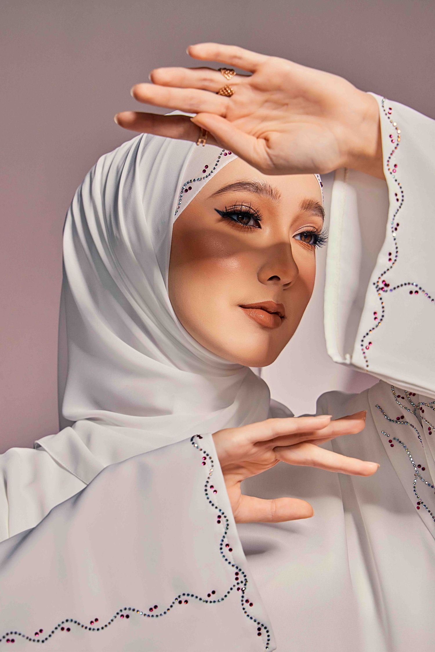Woman in white hijab and embroidered abaya framing her face with both hands in studio portrait.