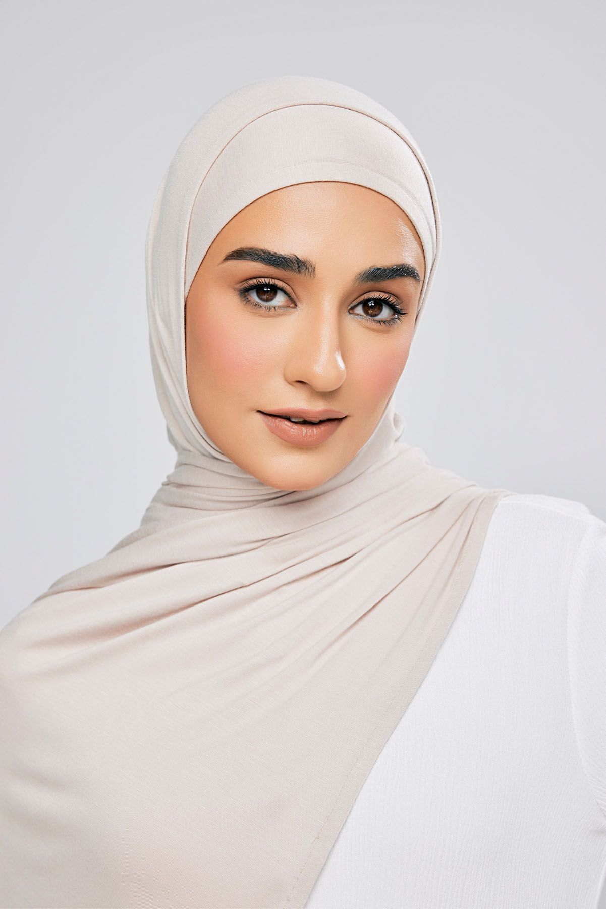 Close-up portrait of a woman in a light beige hijab and white top on a neutral background.