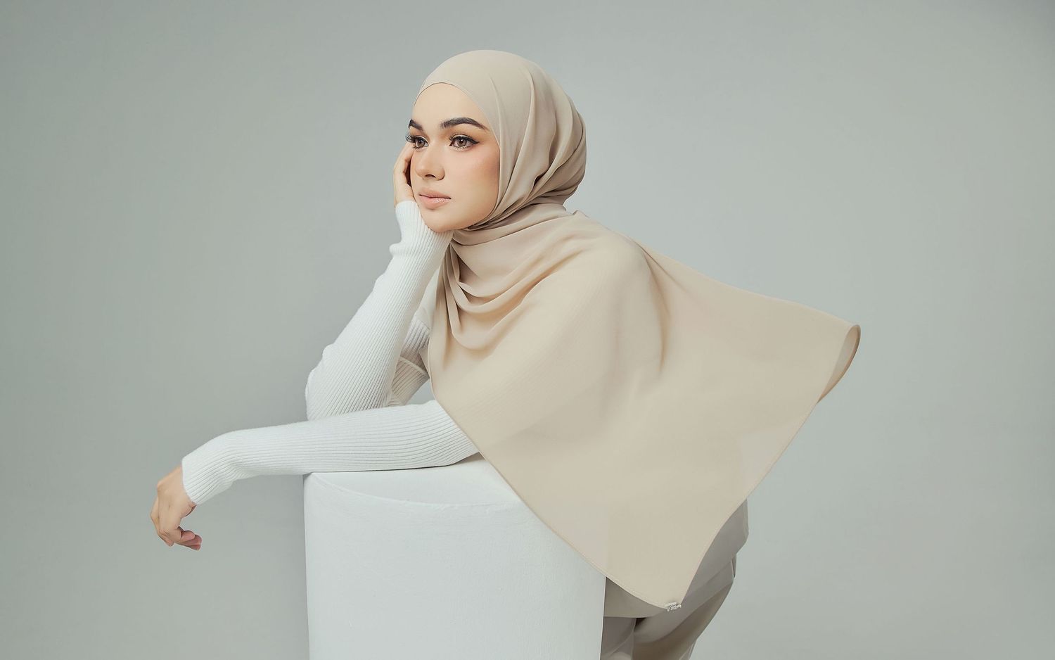 Woman wearing beige chiffon hijab and white long sleeve top posing against grey studio background.