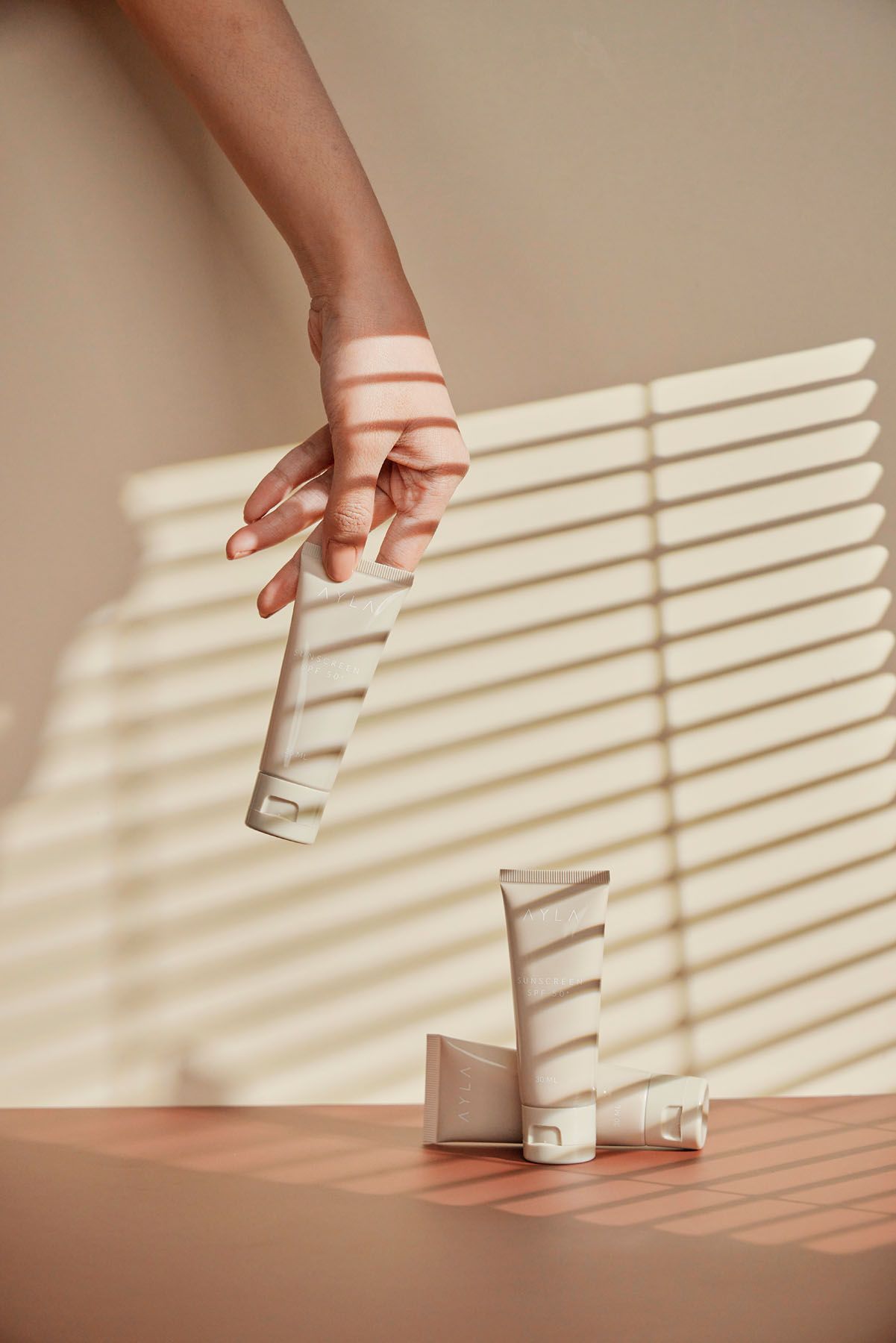 Hand holding skincare tube above two standing cream tubes on terracotta surface with blind shadow pattern.