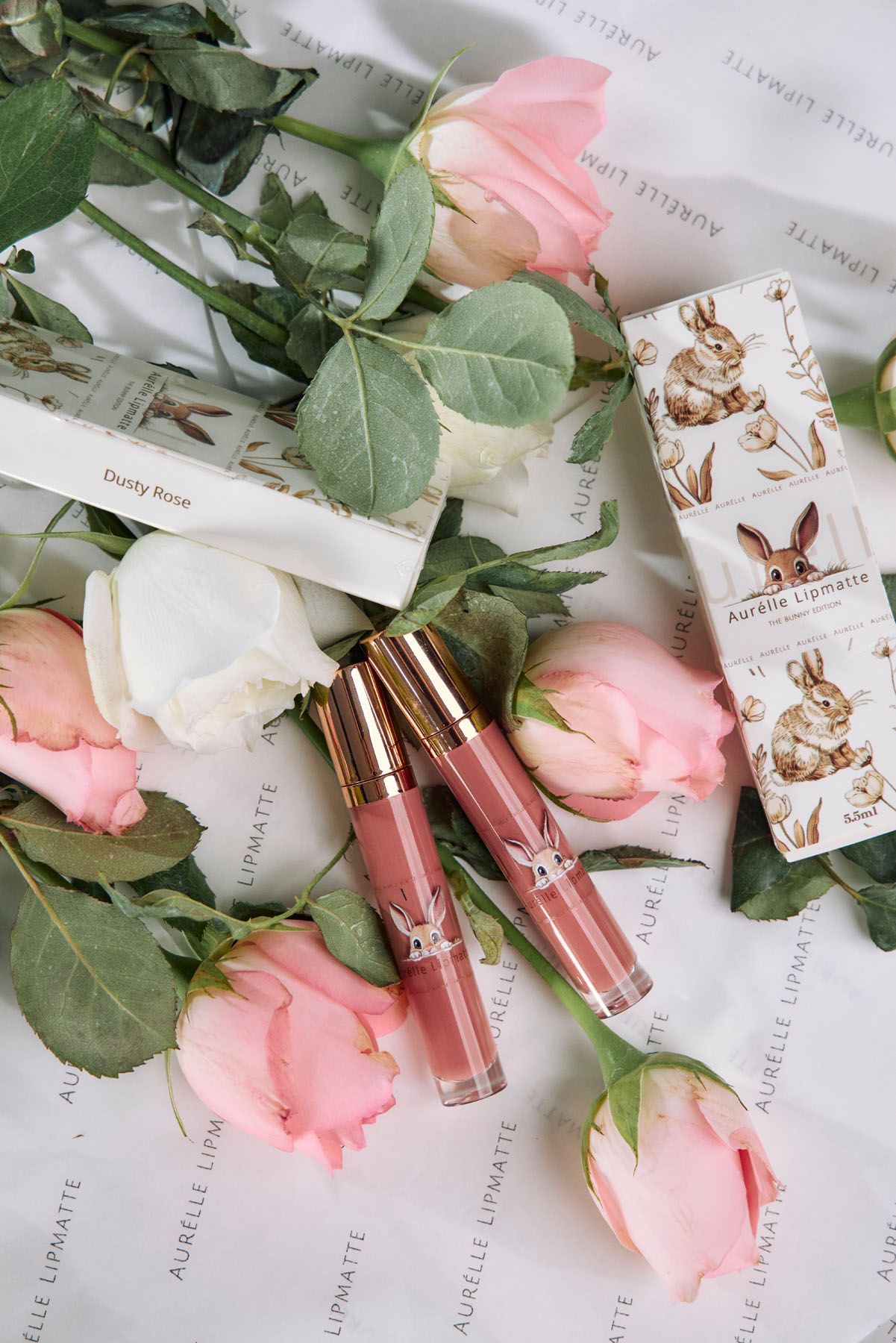 Two Aurelle lip matte tubes in dusty rose shade surrounded by pink and white roses on white fabric.
