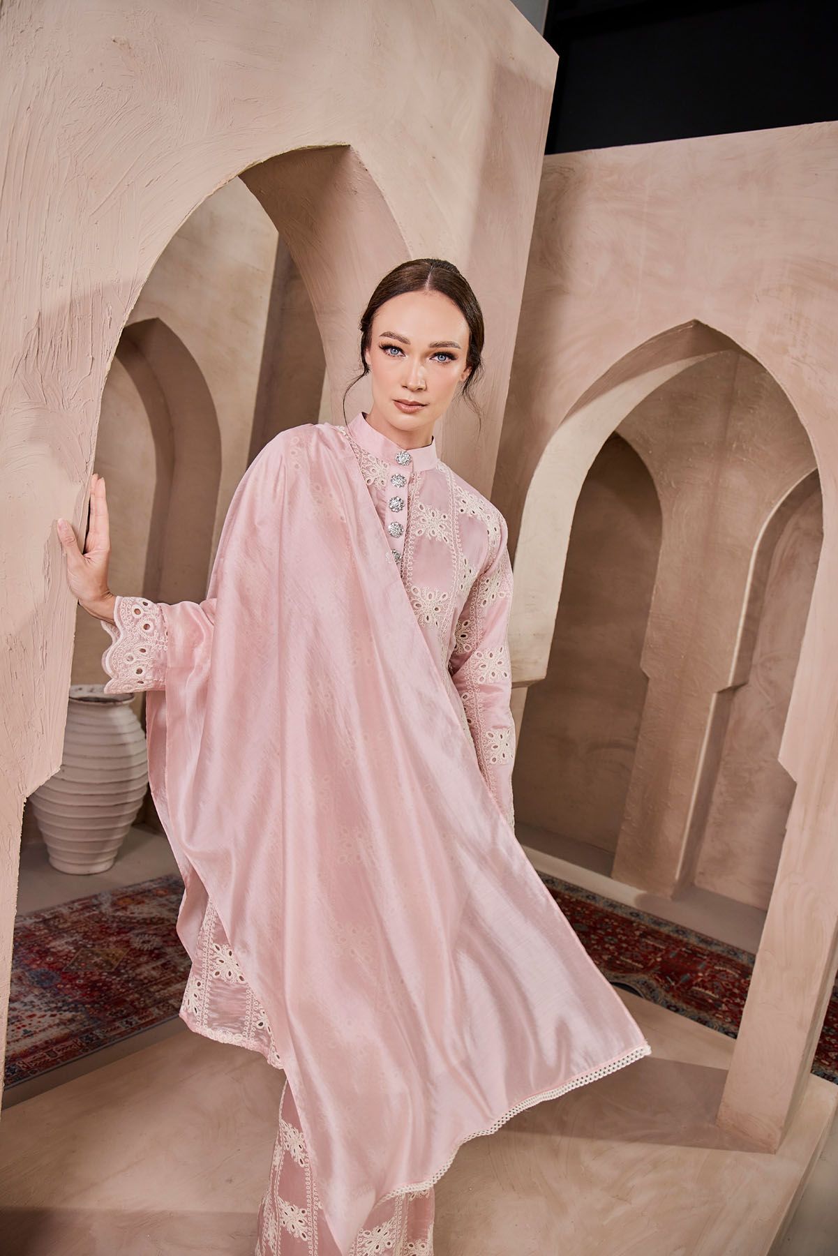 Woman in blush pink embroidered baju kurung with wide sheer shawl poses elegantly.