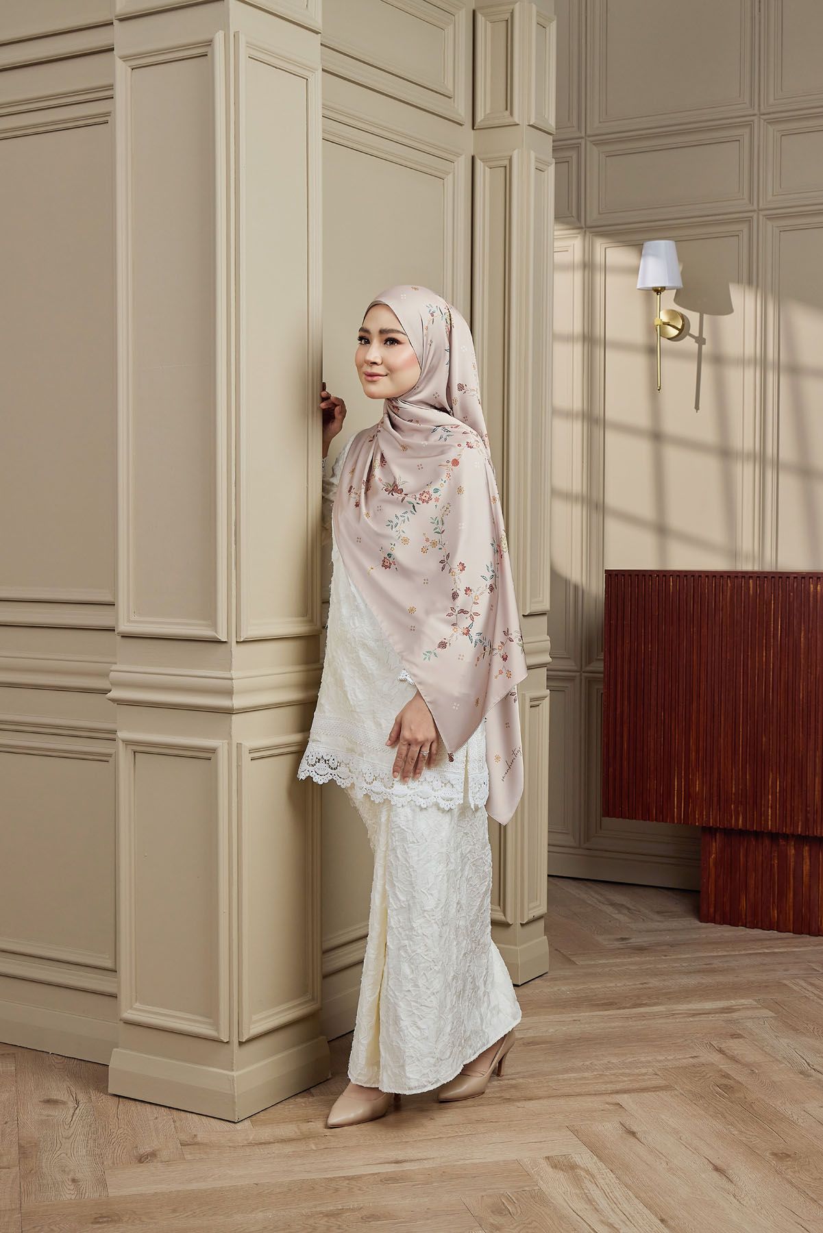 Woman in white lace baju kurung and floral pink hijab posing elegantly against a beige wall.