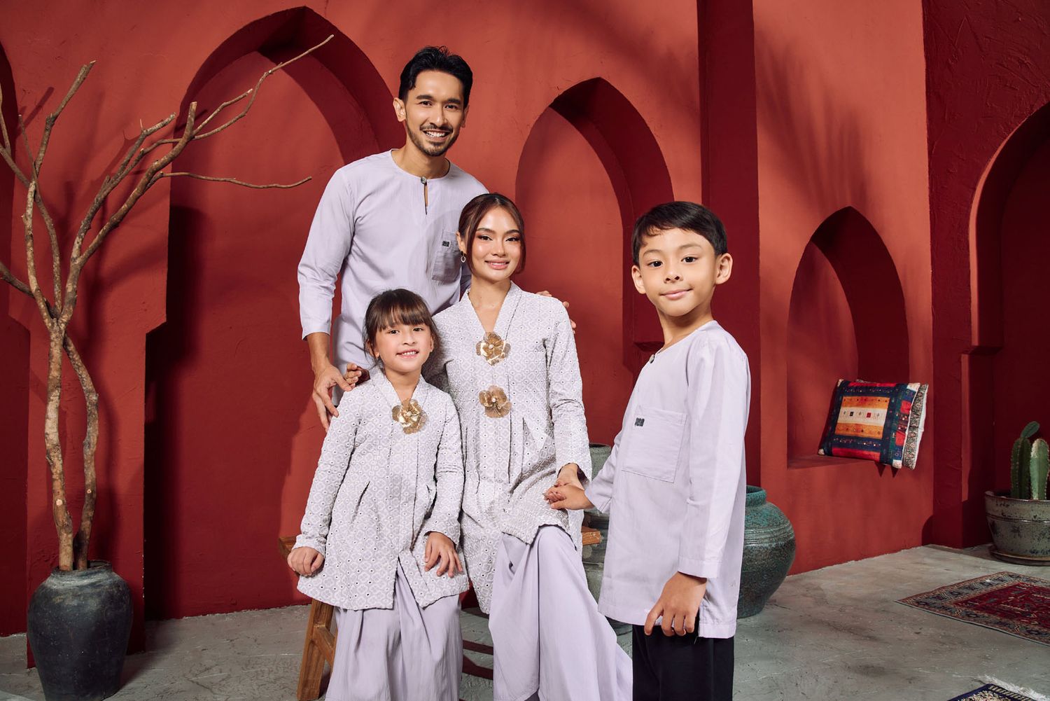 Happy family of four in matching lilac traditional outfits posing together against a vibrant red wall backdrop.