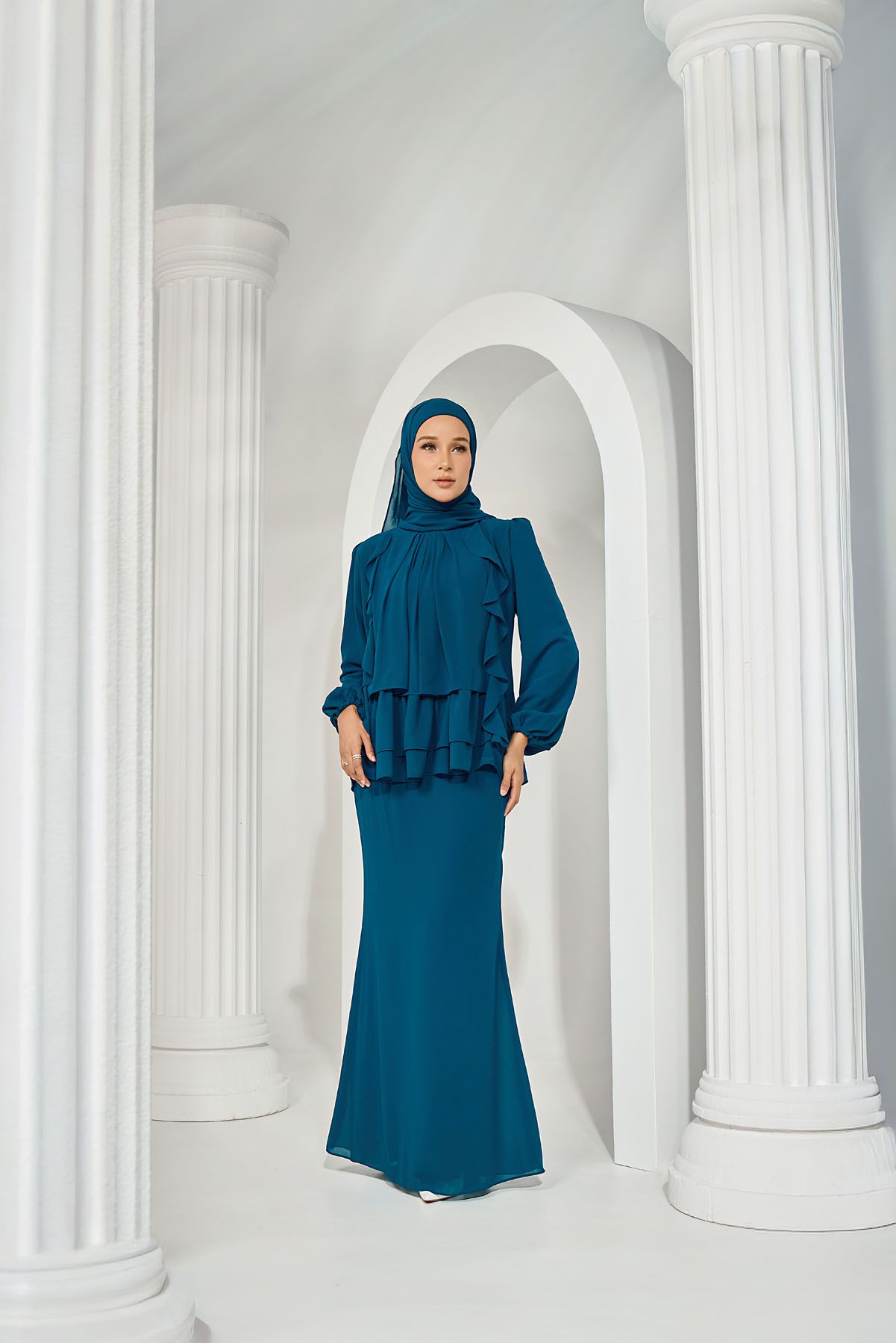 Muslim woman wearing a teal ruffled baju kurung with matching hijab standing between white classical columns.