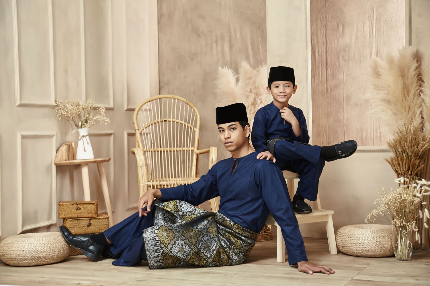 A man and boy in matching navy blue baju melayu pose together in a styled studio setting.