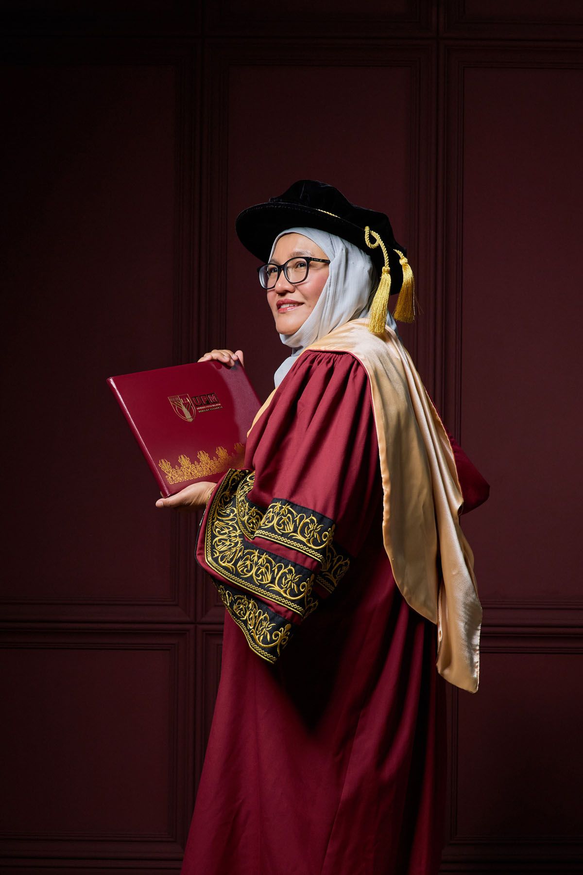 Female graduate in maroon PhD regalia and hijab posing with diploma book at graduation ceremony.