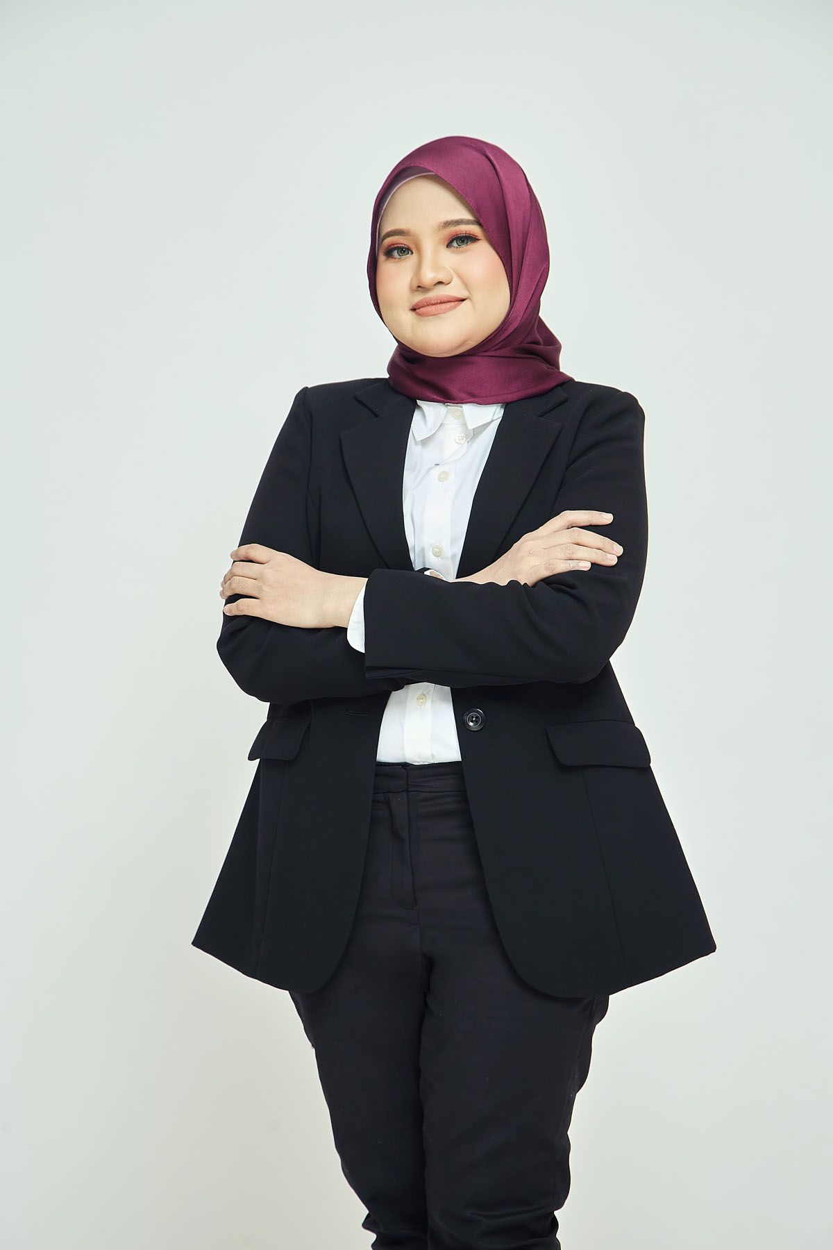 Young Muslim businesswoman in black suit and burgundy hijab posing with arms crossed confidently.