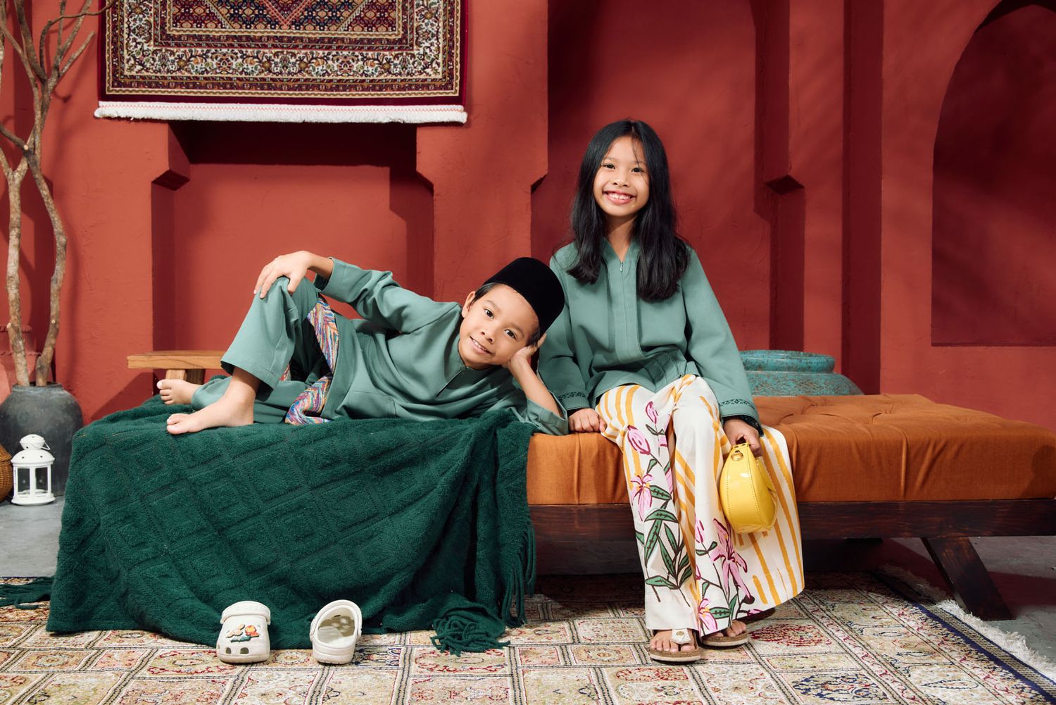 Boy and girl in green baju melayu and baju kurung posing on a daybed in a Moroccan-inspired studio.