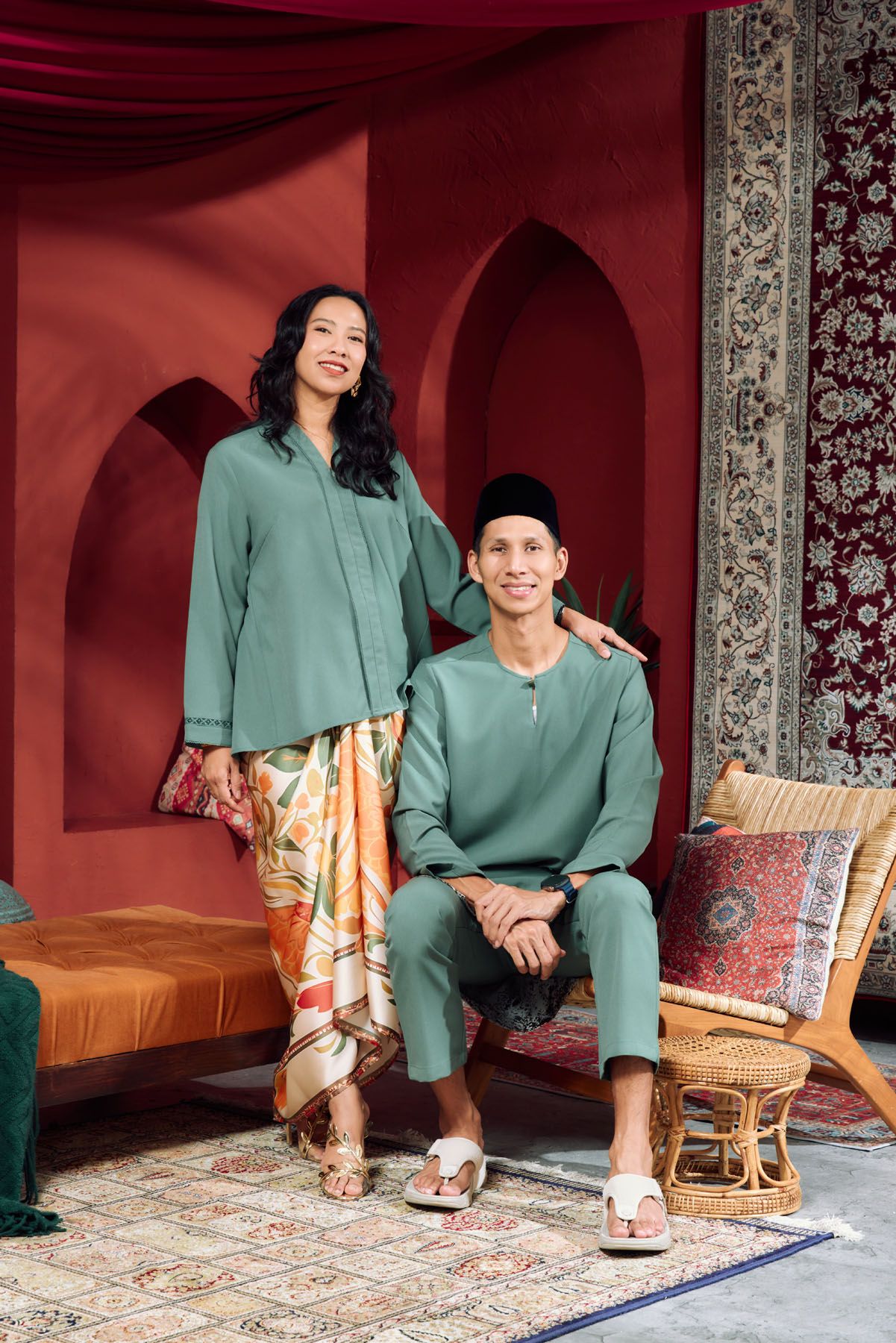 Malaysian couple in matching sage green Baju Melayu and Baju Kurung posing in a red Moroccan-style setting.