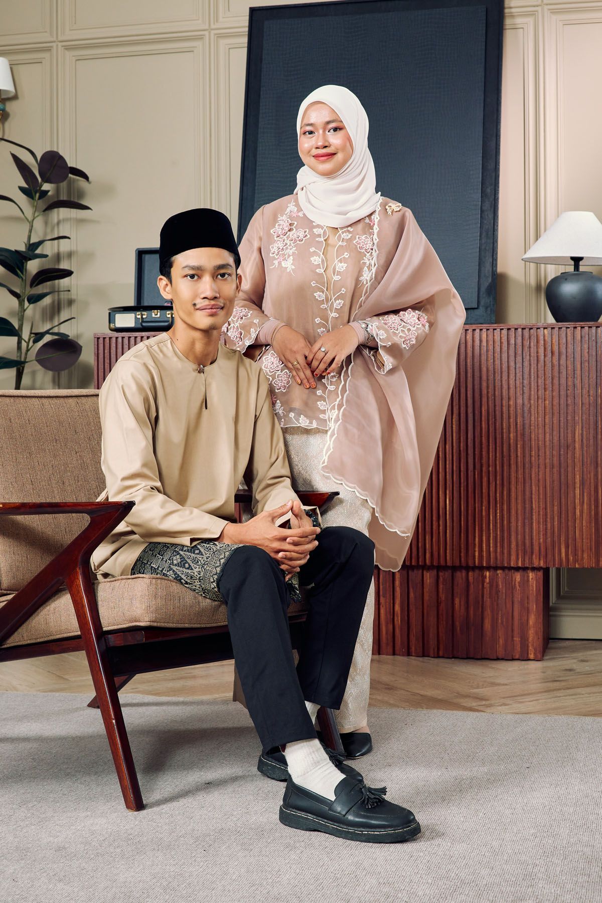 Young Malaysian couple in beige and blush traditional Malay outfits posing in modern interior setting.