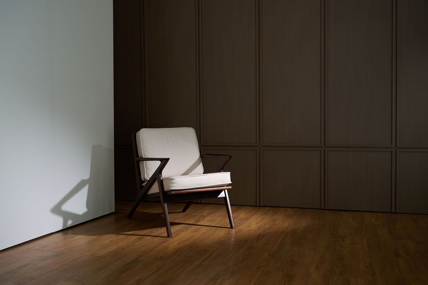Modern white accent chair with wooden Z-frame legs on hardwood floor against dark paneled wall in minimalist room.