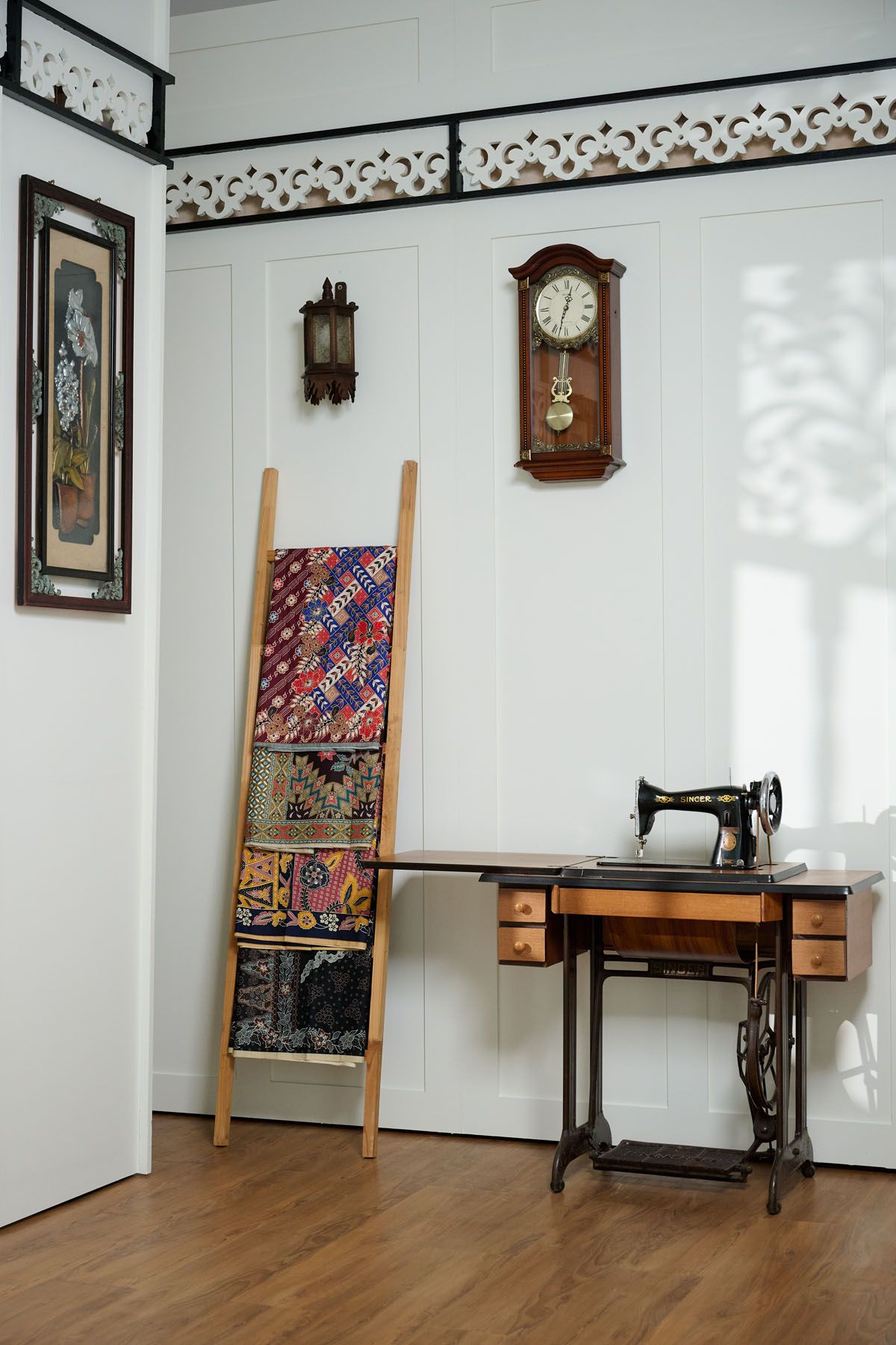 Vintage room with antique sewing machine, wooden ladder displaying colorful quilts, and two wall clocks on white paneled wall.