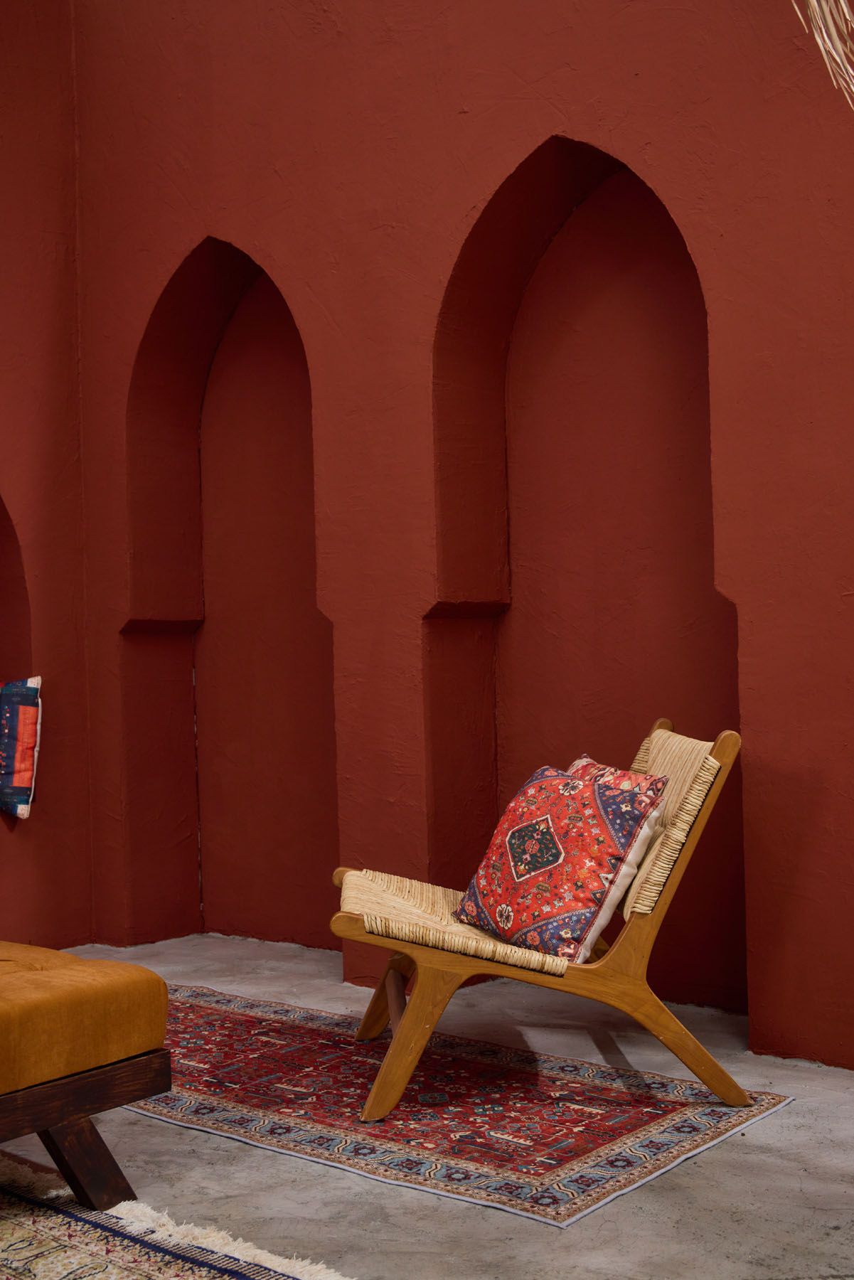 Bohemian rattan lounge chair with Persian rug cushion on red carpet against terracotta arched wall niche backdrop.
