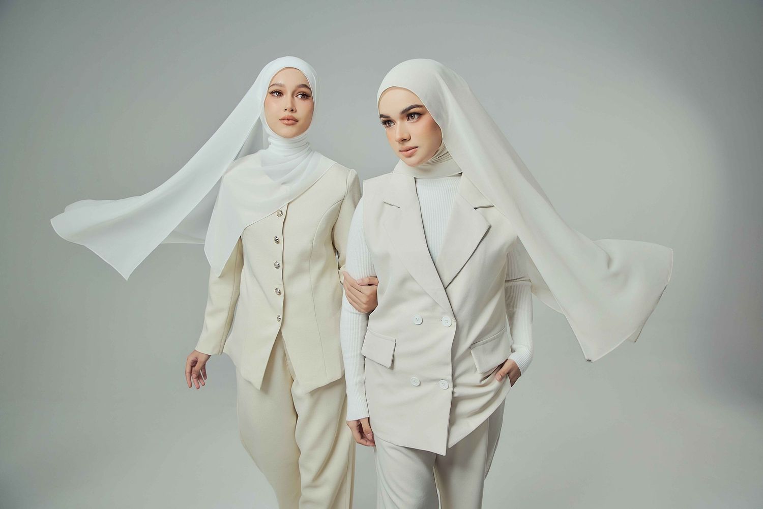 Two women in elegant cream hijab suits with long flowing veils pose against a neutral background.