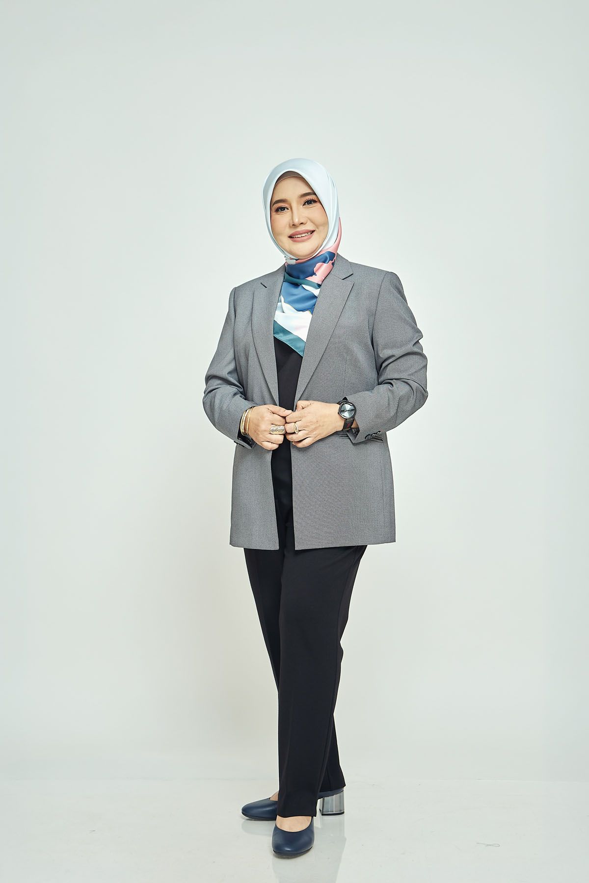 Professional Muslim woman in grey blazer and blue hijab standing confidently on white background.