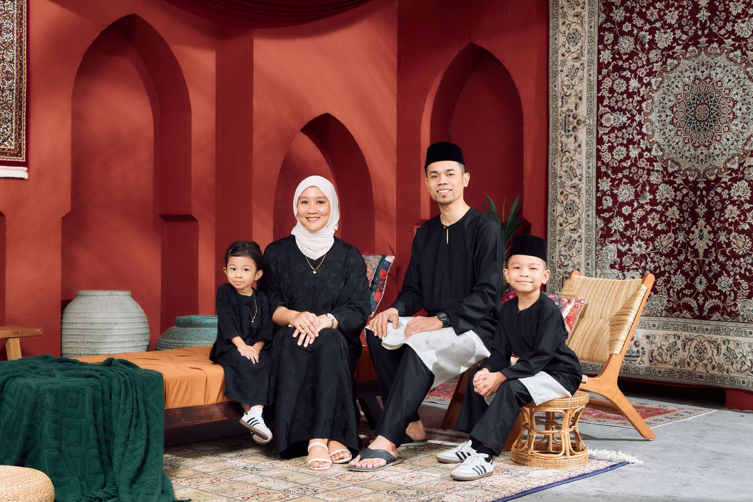Muslim family of four in matching black Baju Melayu and Baju Kurung in red arched setting.
