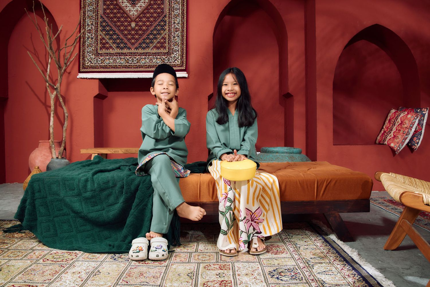 Two children in matching green outfits sitting on a bed in a Moroccan-themed red studio for Hari Raya.