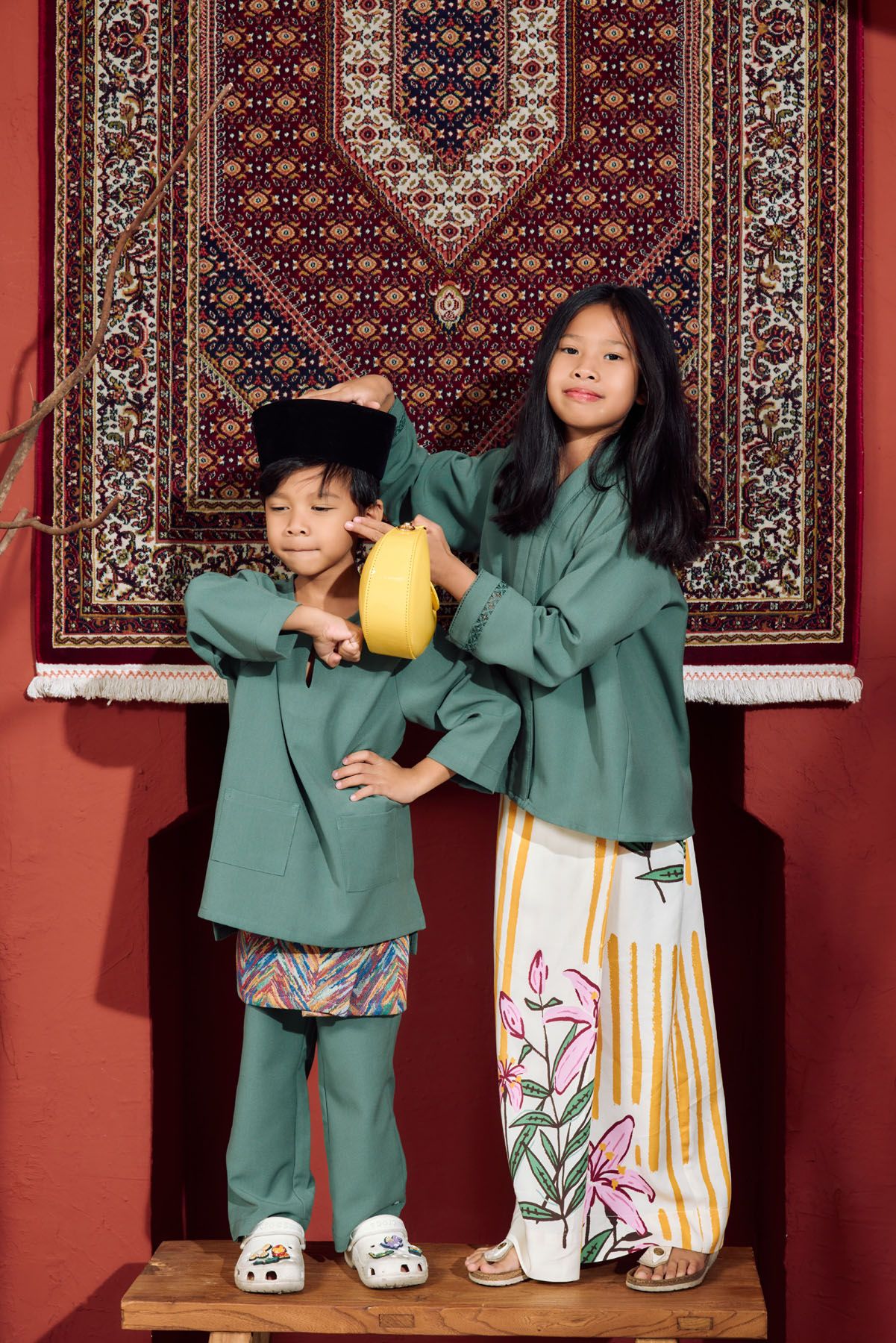 Two children in green traditional Malay outfits standing before a decorative carpet in a red studio.