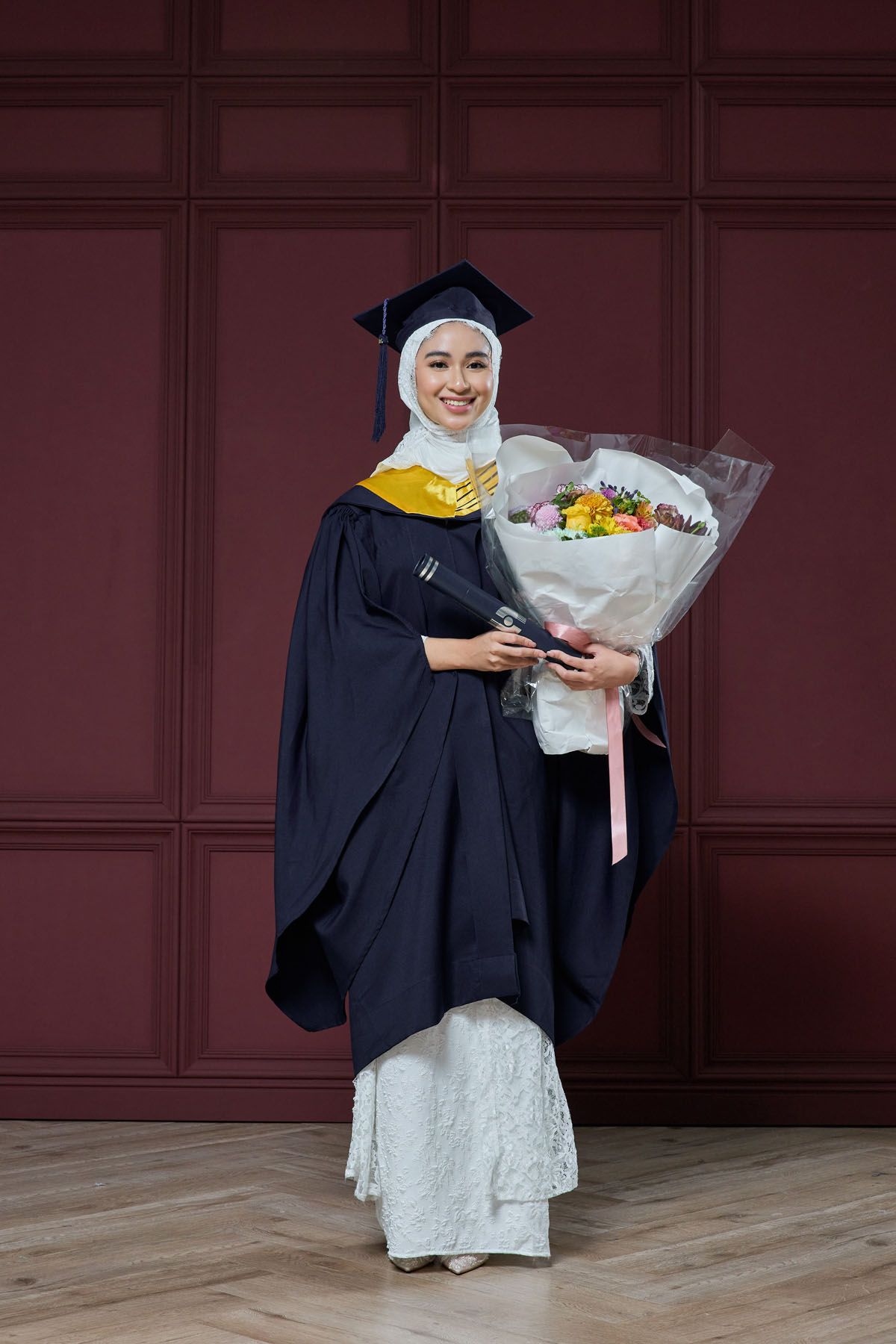 Muslim graduate in navy gown and hijab holding colorful flower bouquet and diploma at ceremony.