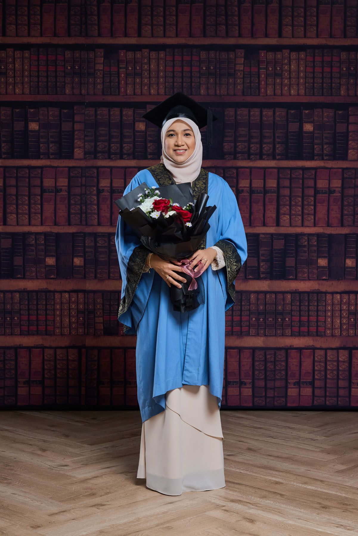 Muslim woman graduate in blue hijab gown holding red and white flower bouquet before bookshelf background.