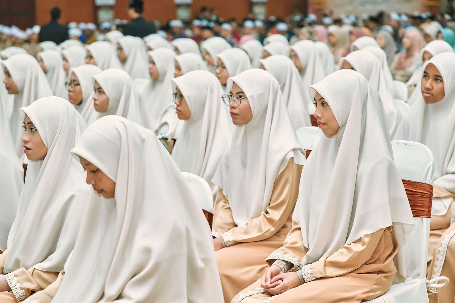 Young Muslim women in white hijabs and beige attire seated attentively at a formal Quran recitation event.