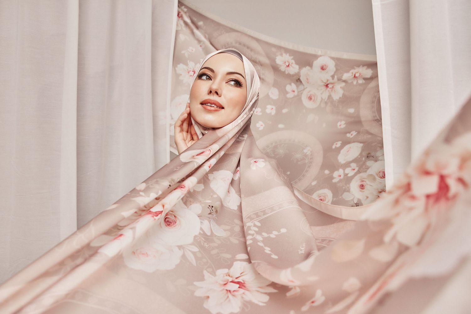 Muslim woman in floral beige hijab peeking through sheer floral curtains in soft natural light.