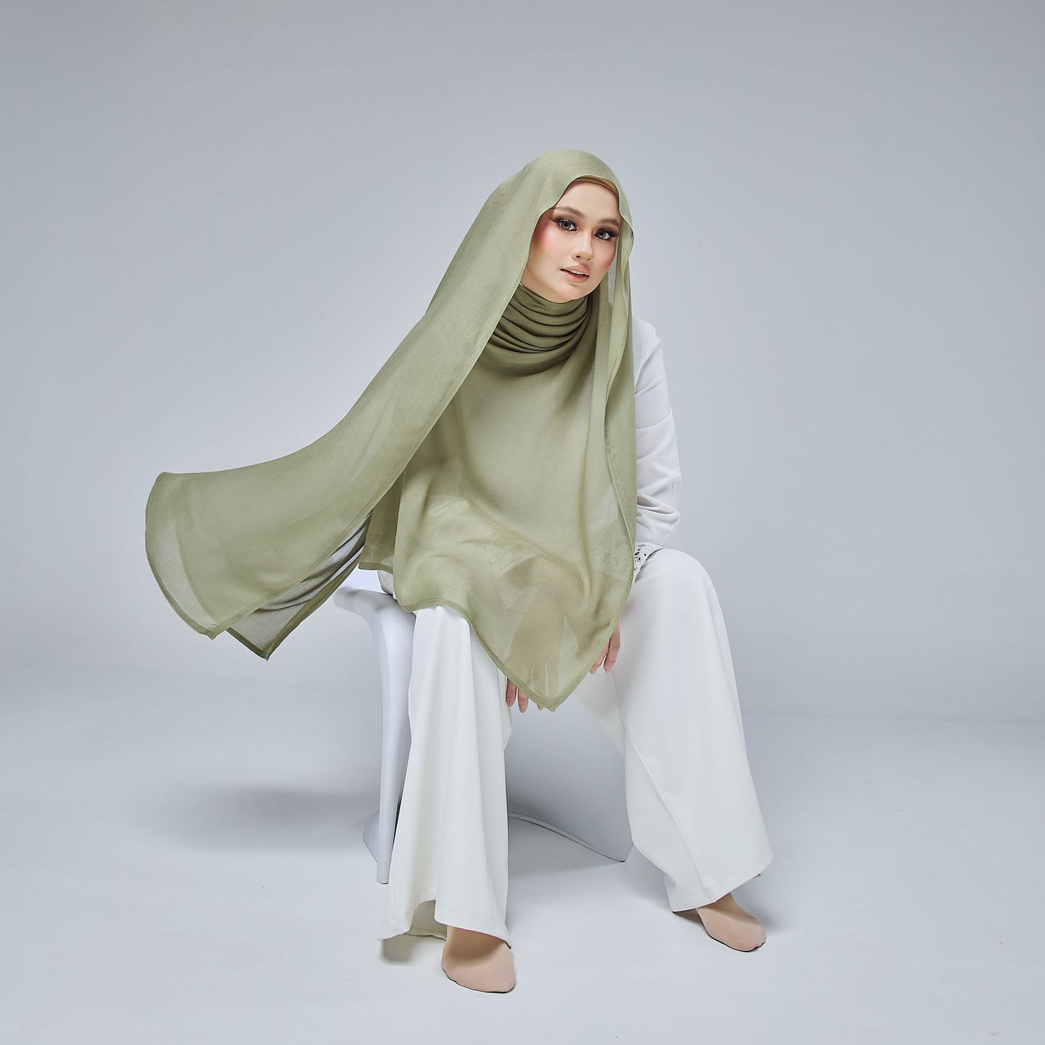 Woman posing on a stool wearing a light olive hijab with fabric flowing in the wind.