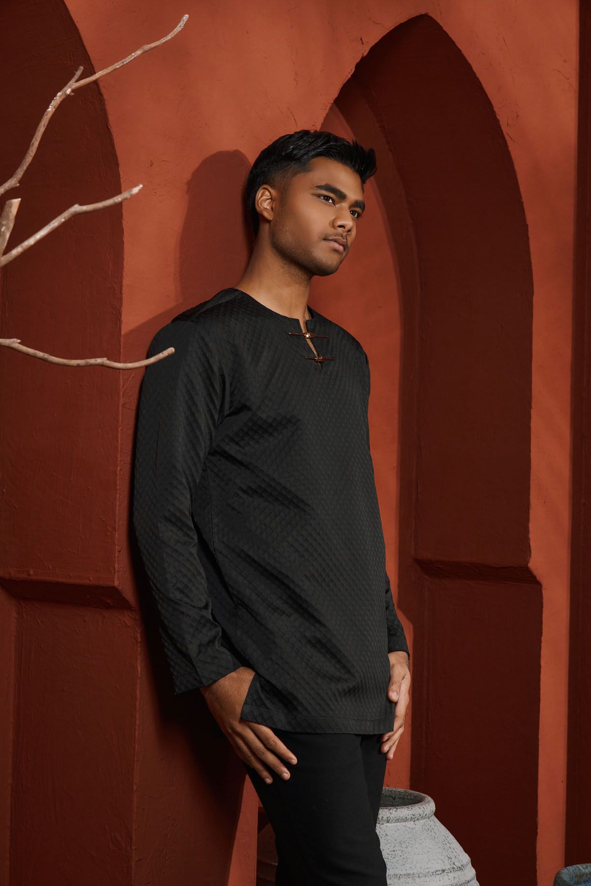 Young man in black quilted baju melayu leaning against a red terracotta wall with arched details.