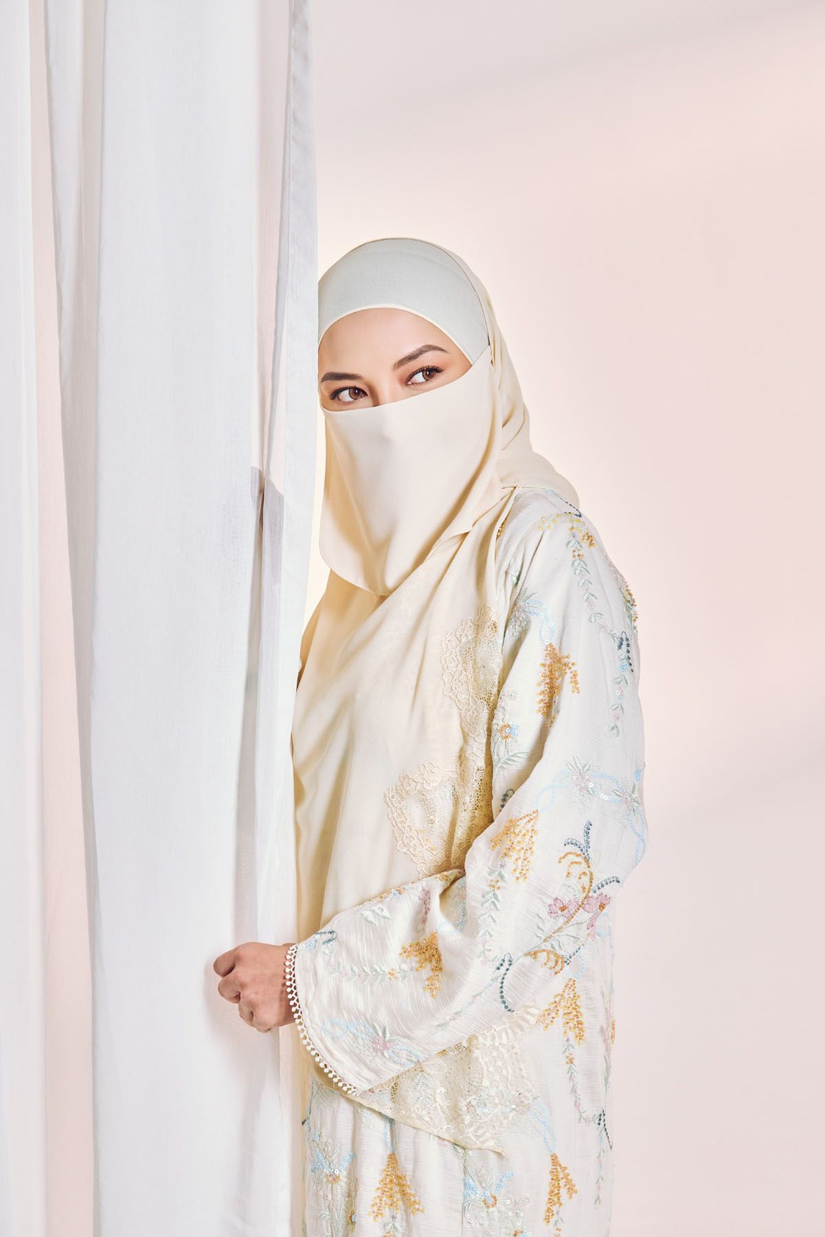 Woman in cream floral abaya and niqab peeks around white curtain.