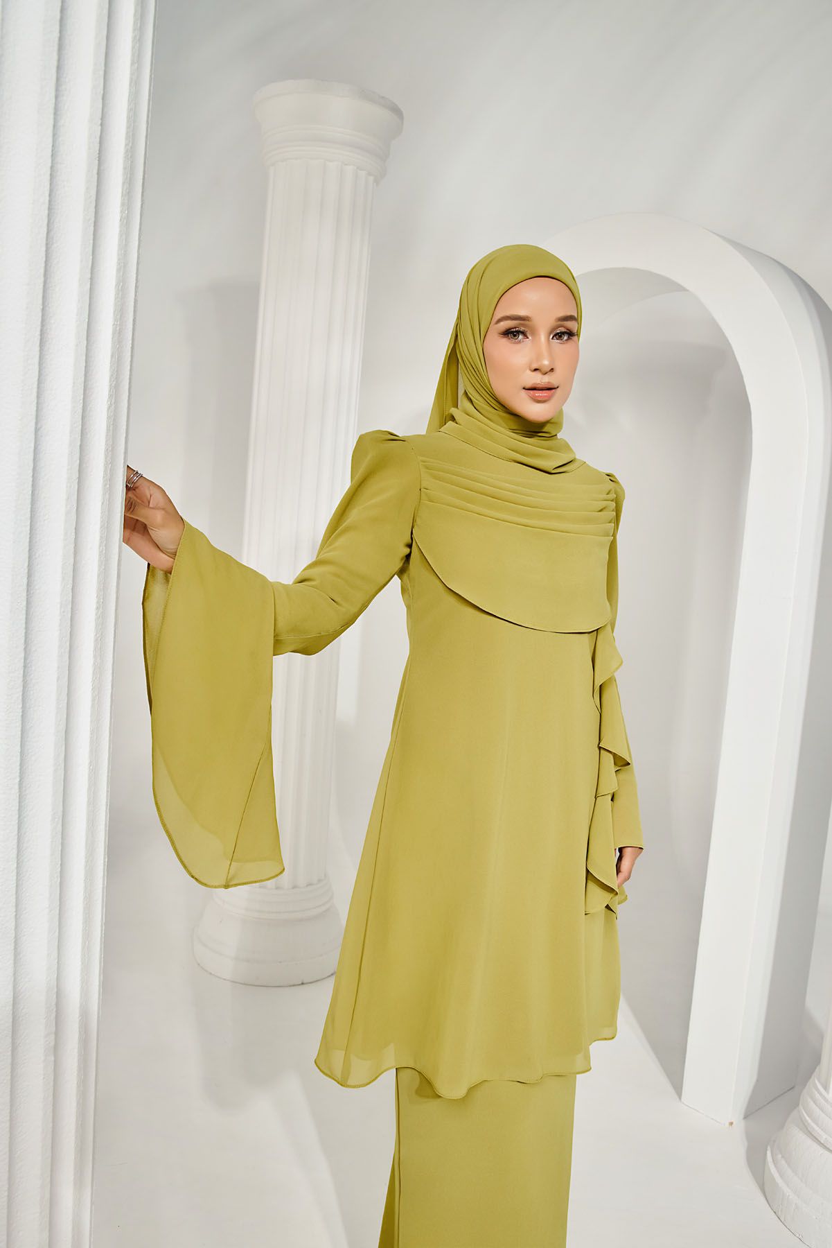 Muslim woman in olive green chiffon modest dress and matching hijab posing beside white architectural columns.