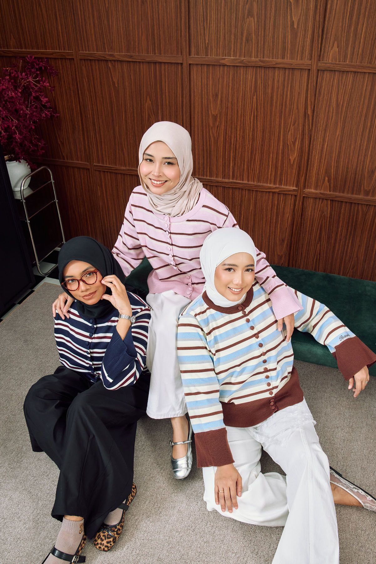 Three smiling women in striped tops and hijabs pose together in a stylish wood-paneled room setting.