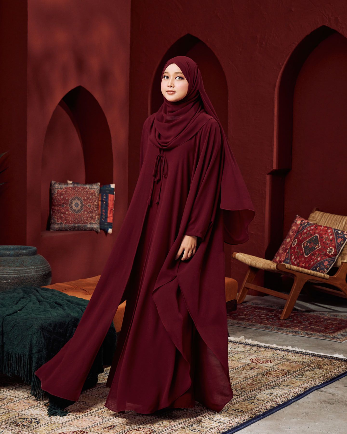 Woman in elegant burgundy abaya and matching hijab poses in a Moroccan-inspired red room with arched alcoves.